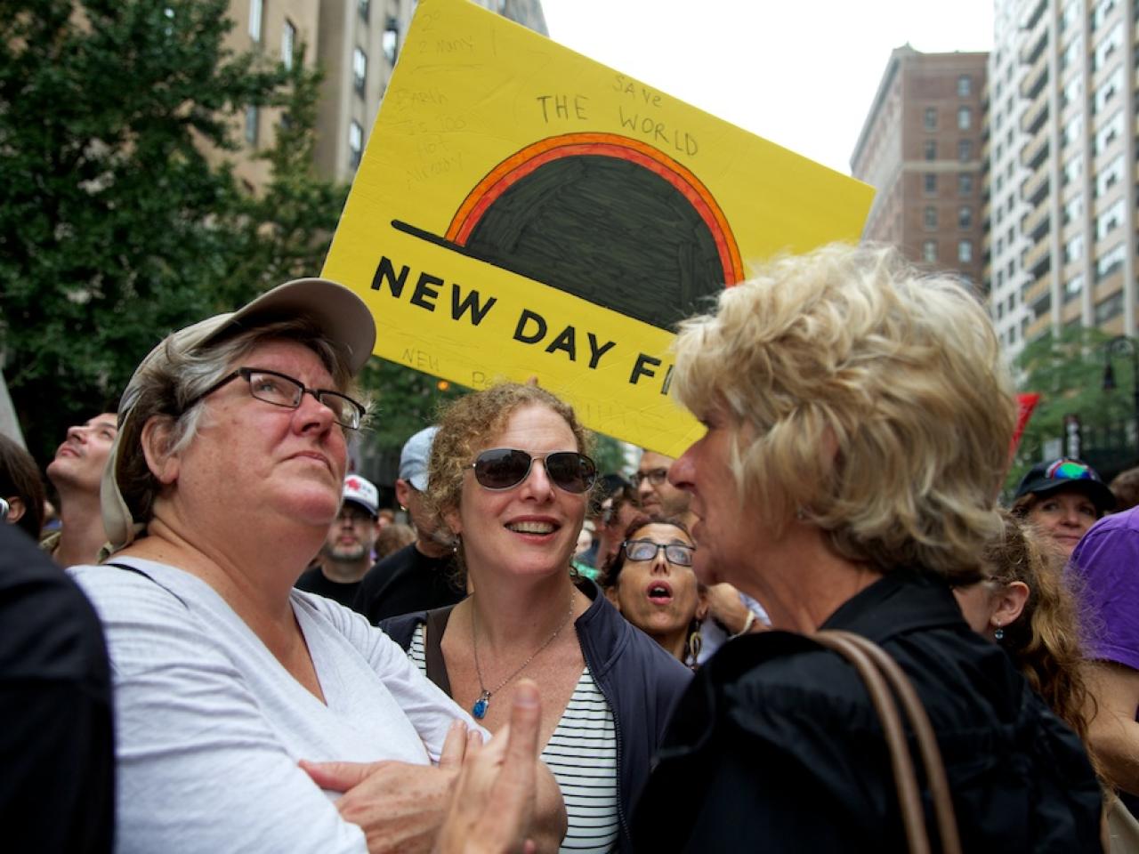 Three people stand in a circle at the People’s Climate March looking in different directions. A large yellow poster with the New Day Films Logo is hoisted behind the middle woman’s head. The poster has various writings on it, including the words “Save the World” written above the logo. People fill the street moving forward behind the three women and tall city buildings rise up on either side of the frame.