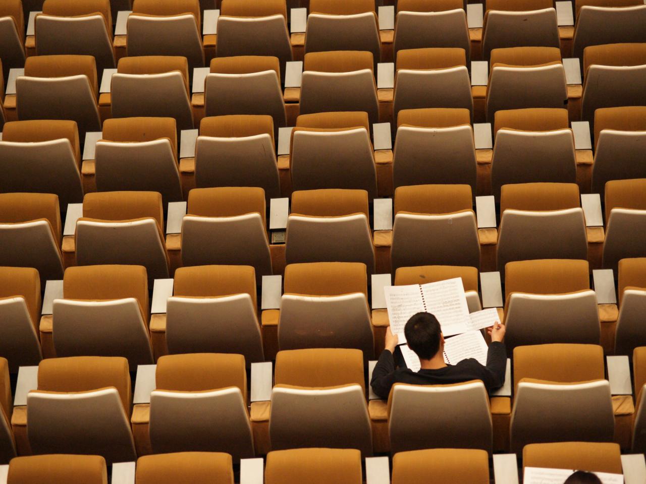 Overhead view of empty, orange seats in an auditorium style classroom. One student in the lower right sits and reads from two open notebooks.