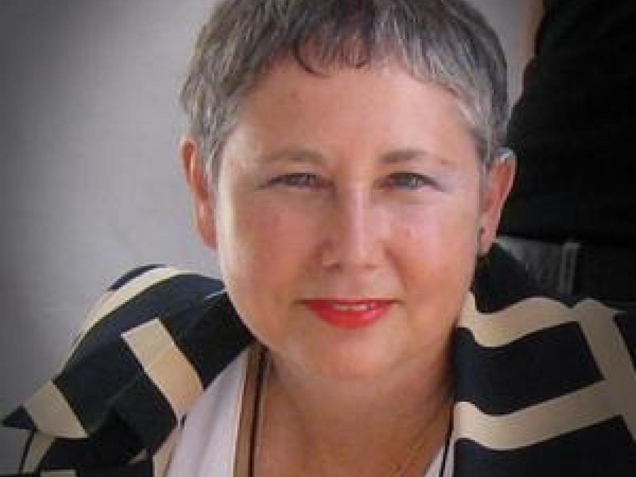 A headshot of New Day filmmaker Sandra Shulberg. A woman with short gray hair and a patterned blazer looks into the camera and smiles. The background is mostly gray with a spotlight behind her head and a stripe of black to the right.