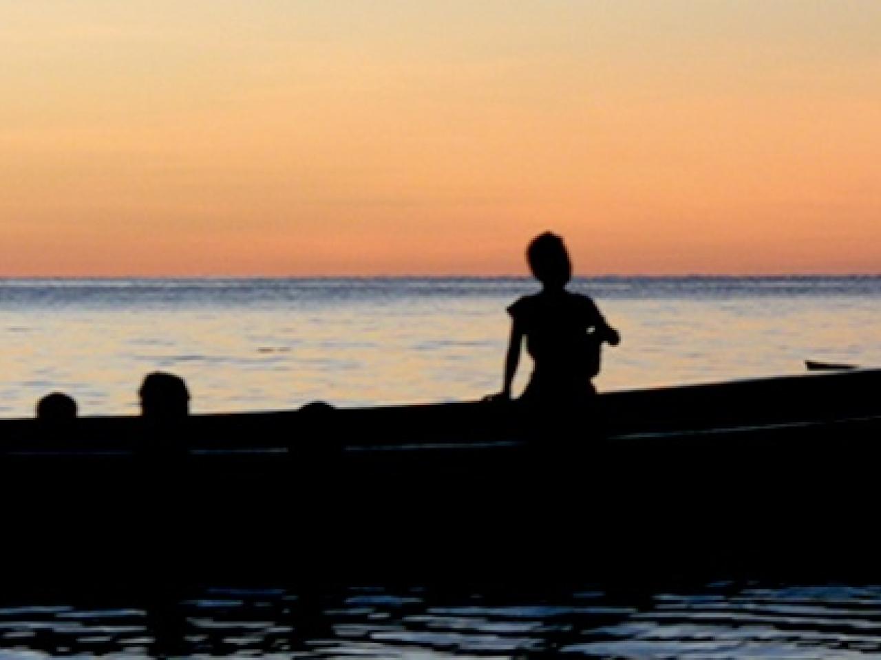 A still from the New Day film Sun Come Up. A silhouette of a large canoe in shiny, calm waters with a deep orange sunset behind. Several people’s heads at different levels show them standing or sitting in the boat. Two people on the right stand at the back of the boat with an oar. A few short buildings disrupt the mostly flat horizon.