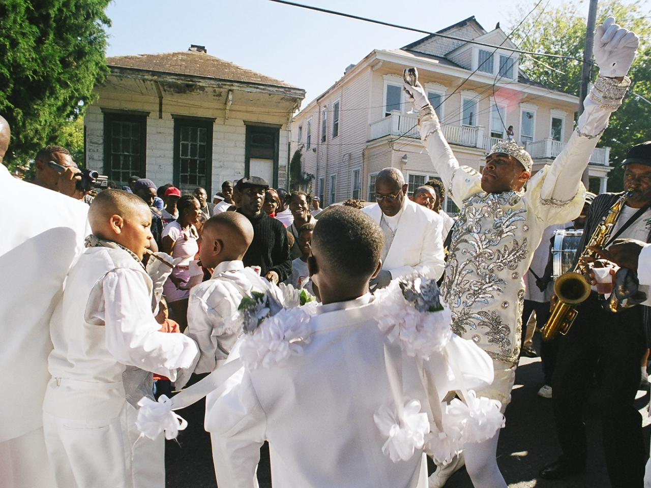 Still from the film “Faubourg Tremé” features dozens of Black community members of all ages gathered at a street parade. They watch a man wearing a sequined white ensemble outfit and white gloves, with both hands raised in the air. He holds a chalice in one hand. Next to him, a man plays the saxophone. In the foreground, several men and boys wear white suits, several adorned with flowery accessories.