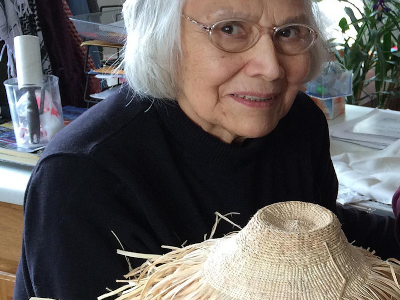 Haida elder and weaver Delores Churchill smiles at the camera. She is wearing glasses and a black top. She holds up a weaving work in progress with the unfinished ends splaying out.