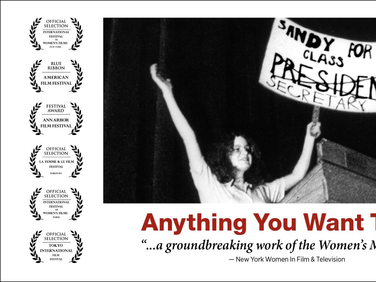 In this black and white still from the film, a young woman triumphantly holds up a sign that reads “Sandy For Class President.” The word ‘President’ is crossed out and underneath is written “Secretary”. She’s onstage at a lectern. Film title "Anything You Want To Be" and festival award laurels.
