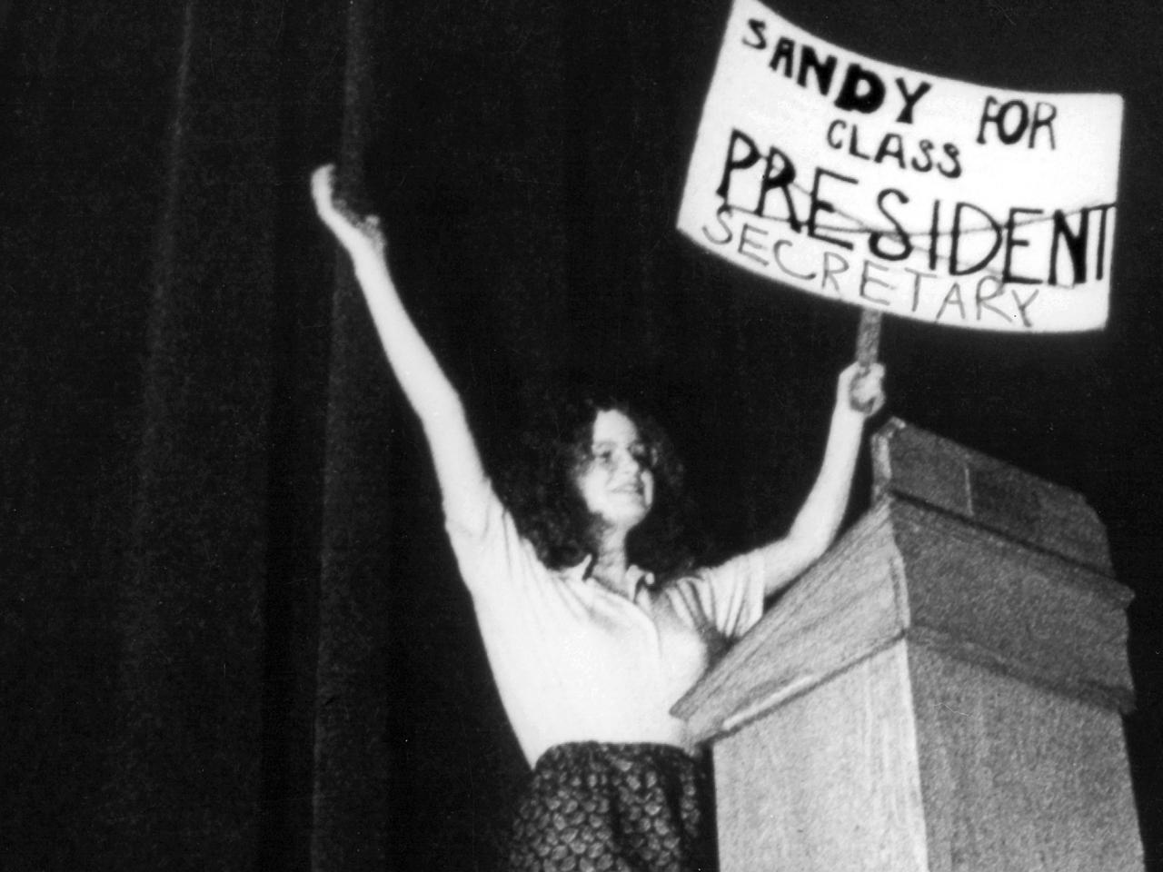 In this black and white still from the film, a young woman triumphantly holds up a sign that reads “Sandy For Class President.” The word ‘President’ is crossed out and underneath is written “Secretary”. She’s onstage at a lectern.