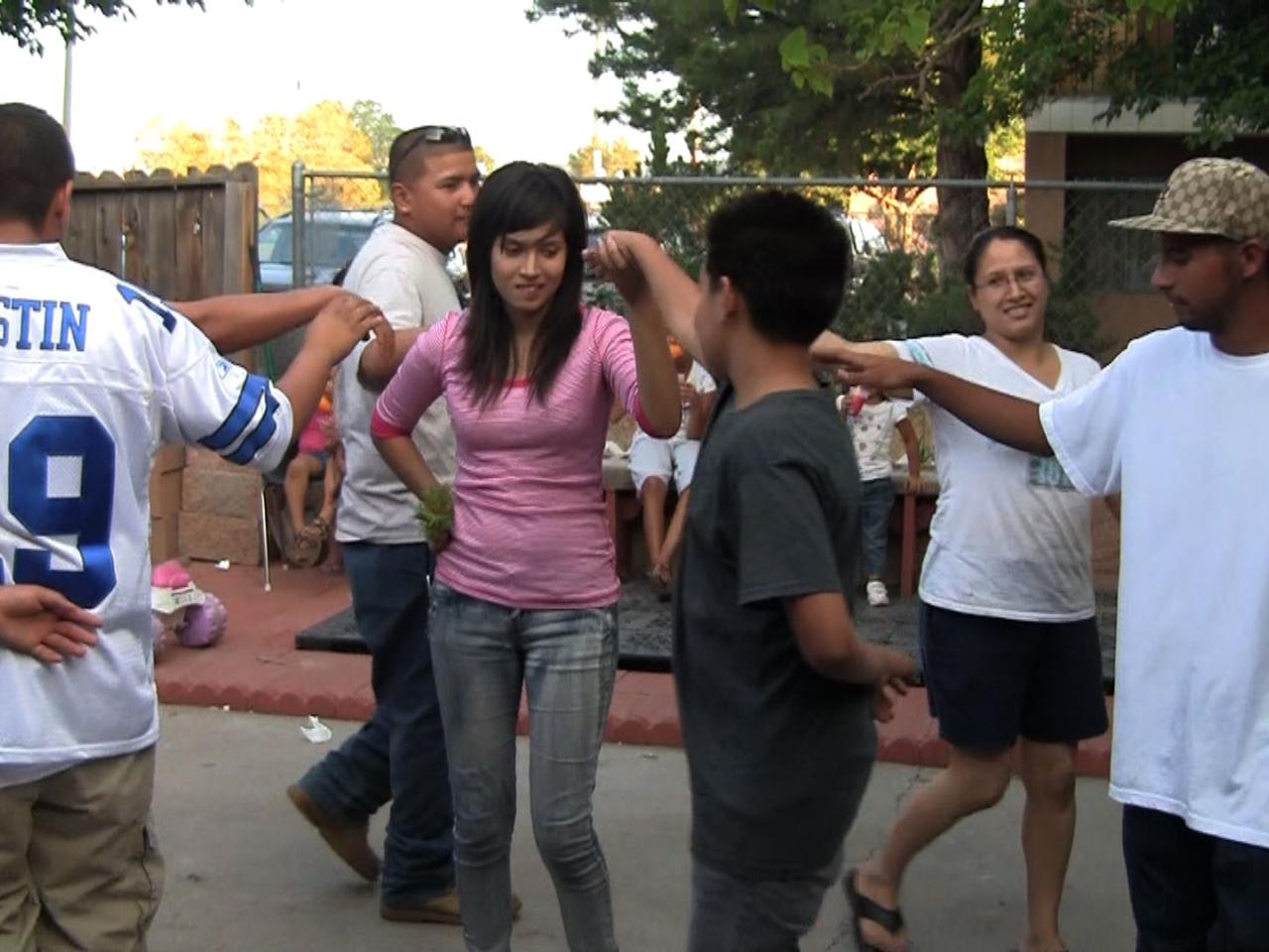 A group of seven people, mostly young, are dancing. They hold one arm outstretched and the other behind their backs. In the center, a girl and a boy are holding hands.