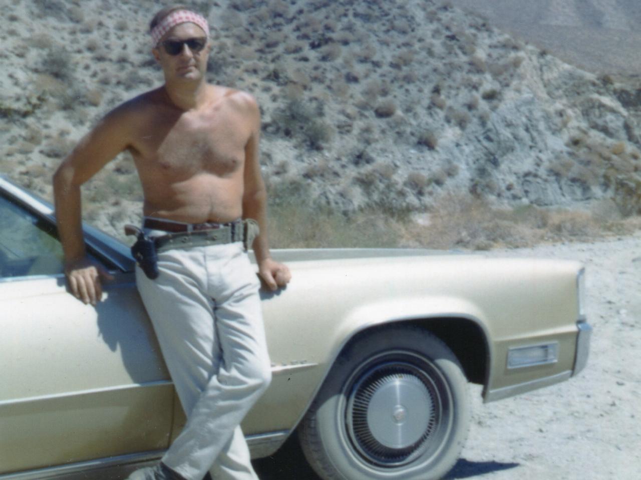 A still from the New Day film The Self-Made Man. An old color photo with off-white edges like a polaroid. Bob Stern, a shirtless man in well worn jeans and sunglasses leans against the side of a tan car from the 70s. The car is on a dusty, dirt road and there are mountains in the background filling the rest of the frame.