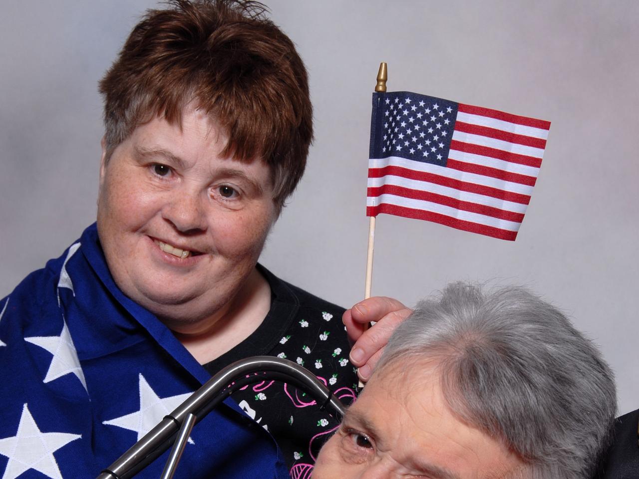 A still from the New Day Film Body & Soul: Diana and Kathy. Two women lean into each other, with the red haired woman’s cheek pressing against the side of the gray haired woman’s forehead. The red haired woman is wearing a blue shirt with white stars and the gray haired woman wears a red shirt. They both look towards the camera and smile.