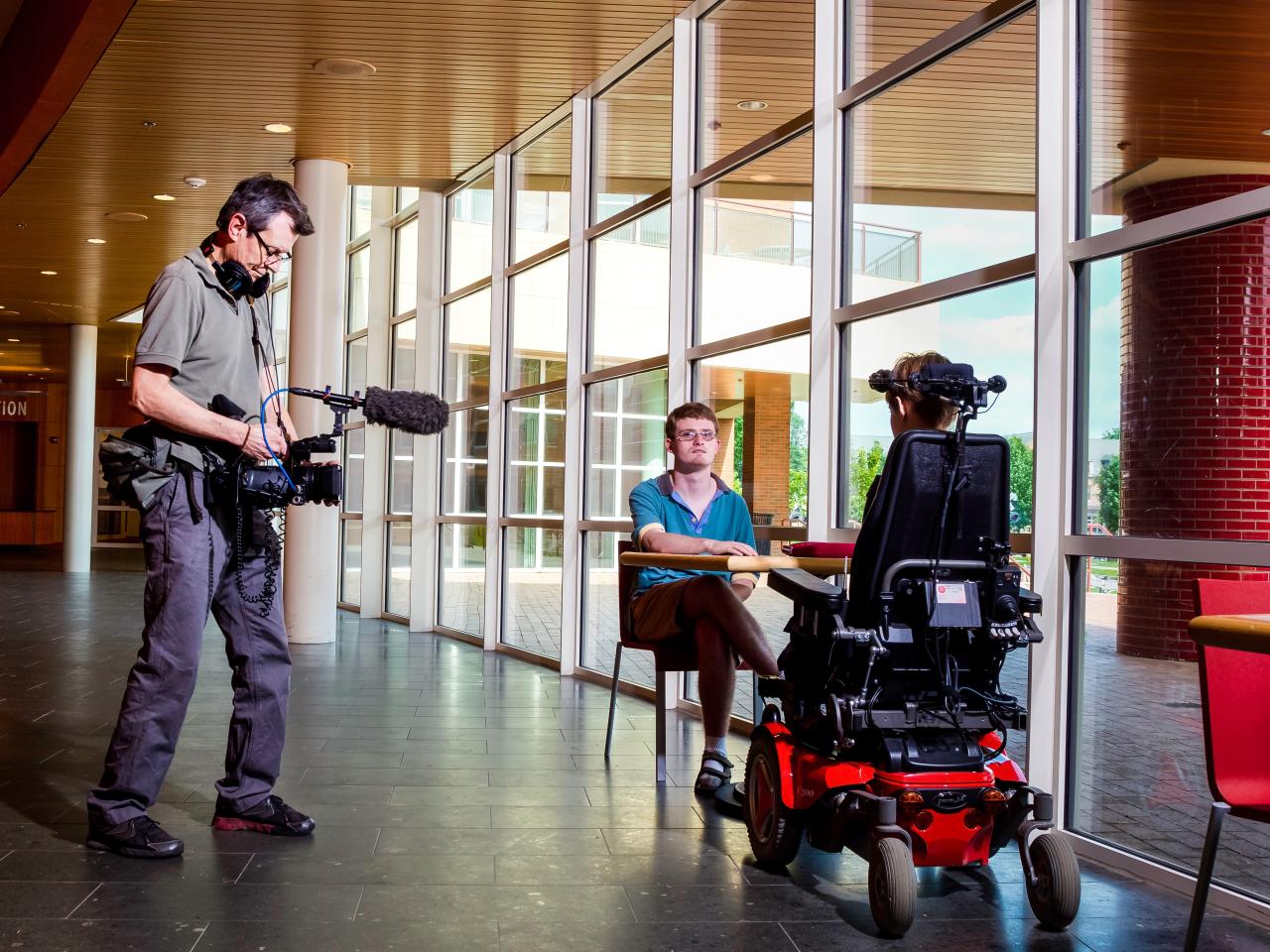 Filmmaker Robert Rooy is standing in a building and is pointing a video camera at two people sitting at a table. One of the people is in a wheel chair. 