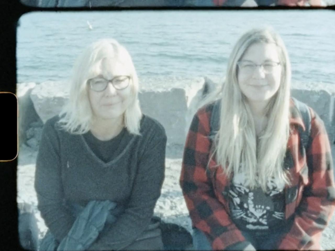 A woman with medium blond hair with her daughter with long blond hair sitting in front of ocean waves