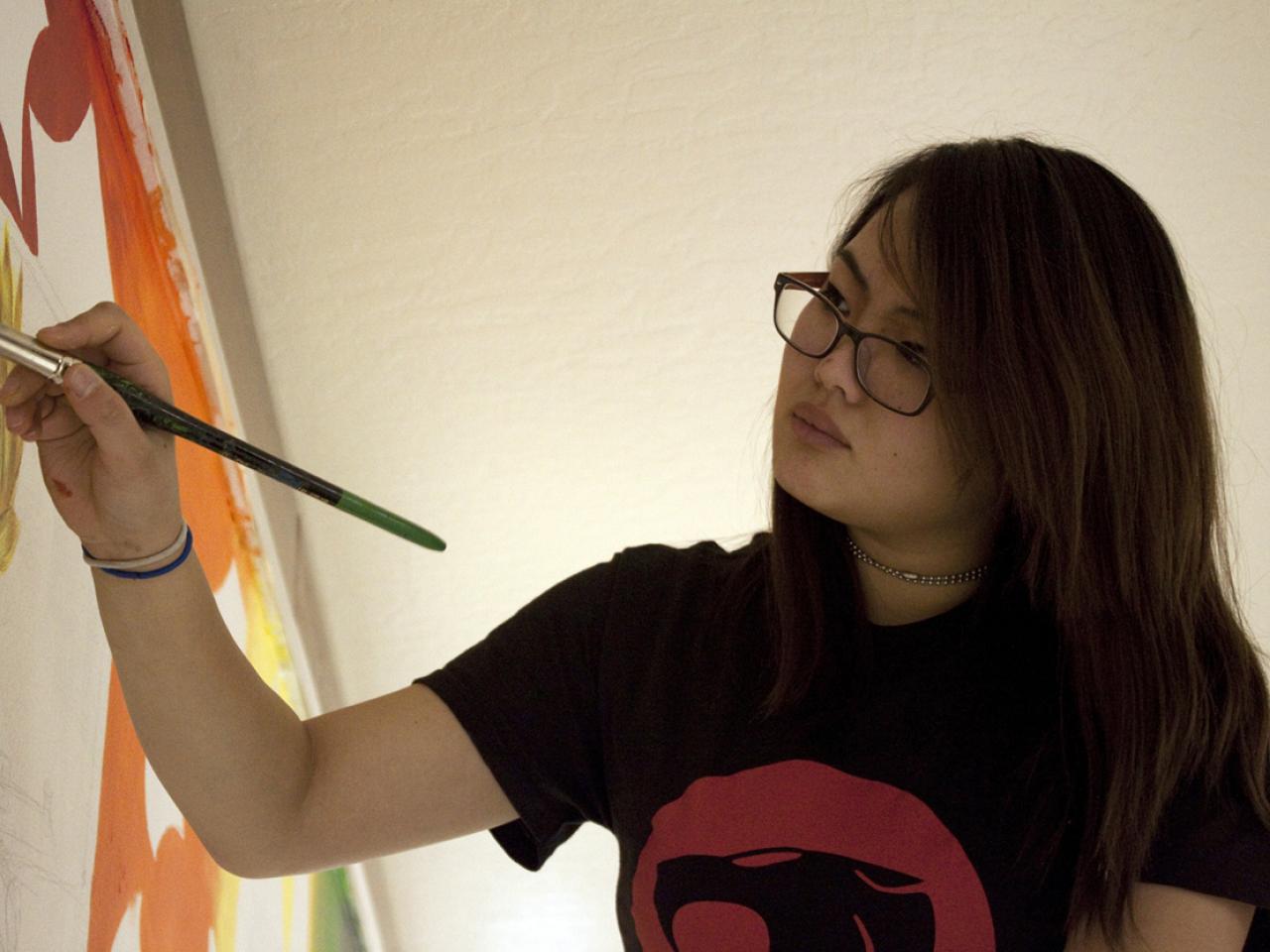 A still from the film Making Noise in Silence shows a Korean teenager painting on a large canvas on an easel.