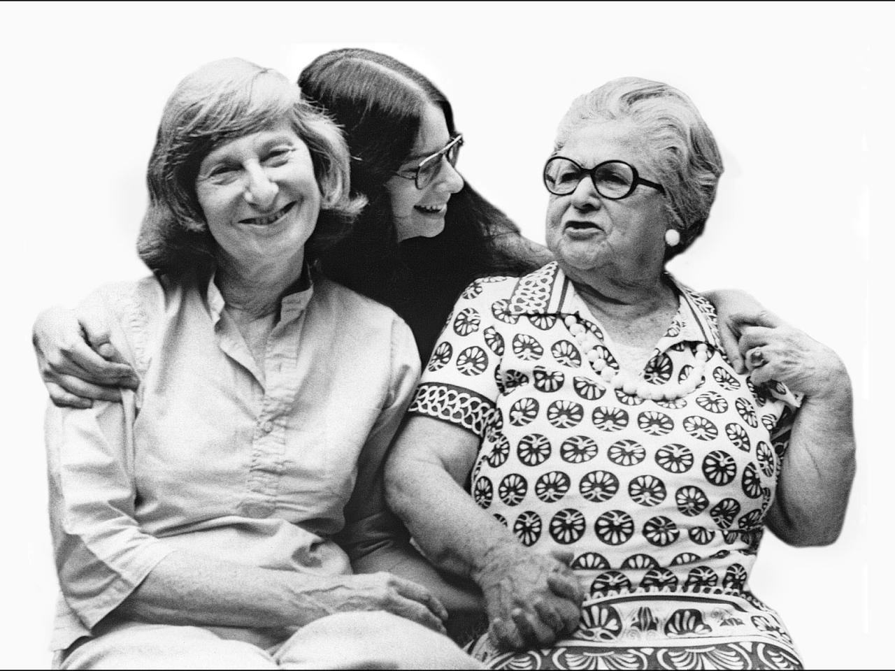 A black and white still from the New Day film, Nana, Mom and Me. A young woman stands behind her mother and grandmother with her arms around them. She glances affectionately towards her grandmother. The grandmother holds her granddaughter’s hand on her shoulder and her daughter's hand on her lap. Beside her, the mother smiles towards the camera.