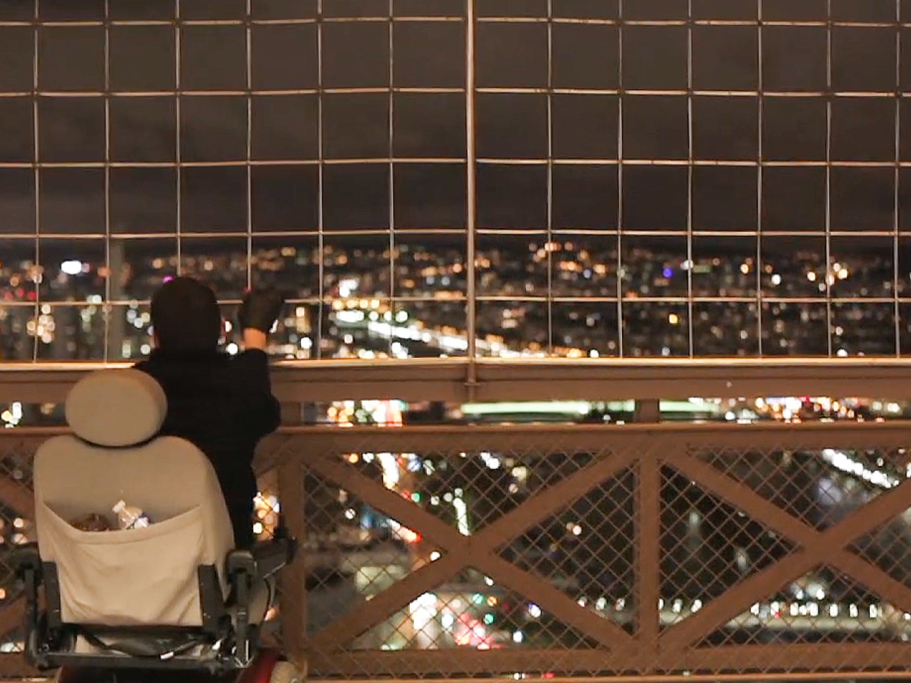 A person in a motorized wheelchair gazes through a metal fence at a brightly lit city skyline at night, in a still from Reid Davenport’s film Wheelchair Diaries
