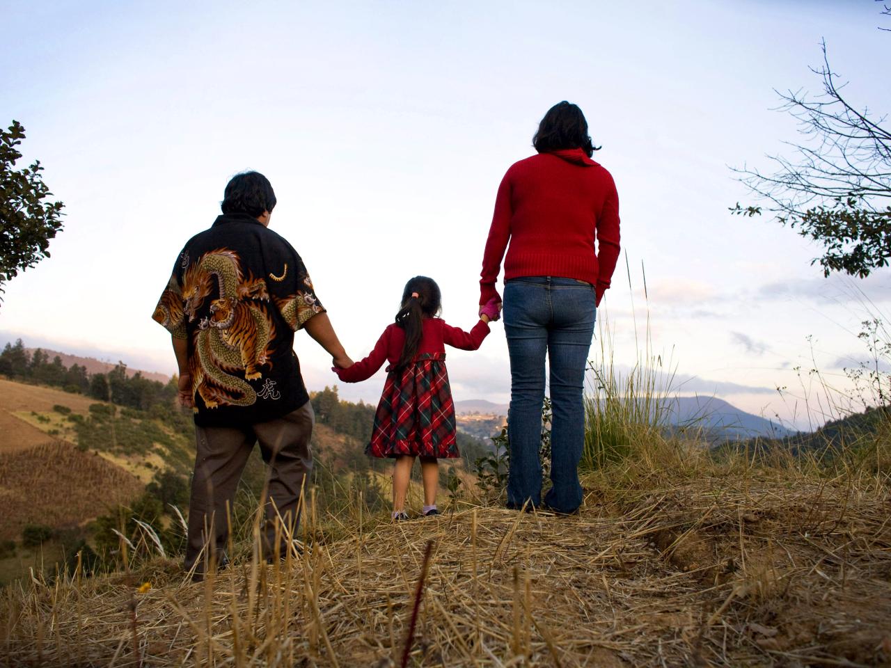 A still from the New Day film Sin Pais. The backs of three family members holding hands, a small child in the middle. They walk on a sandy path with tall grass on either side.