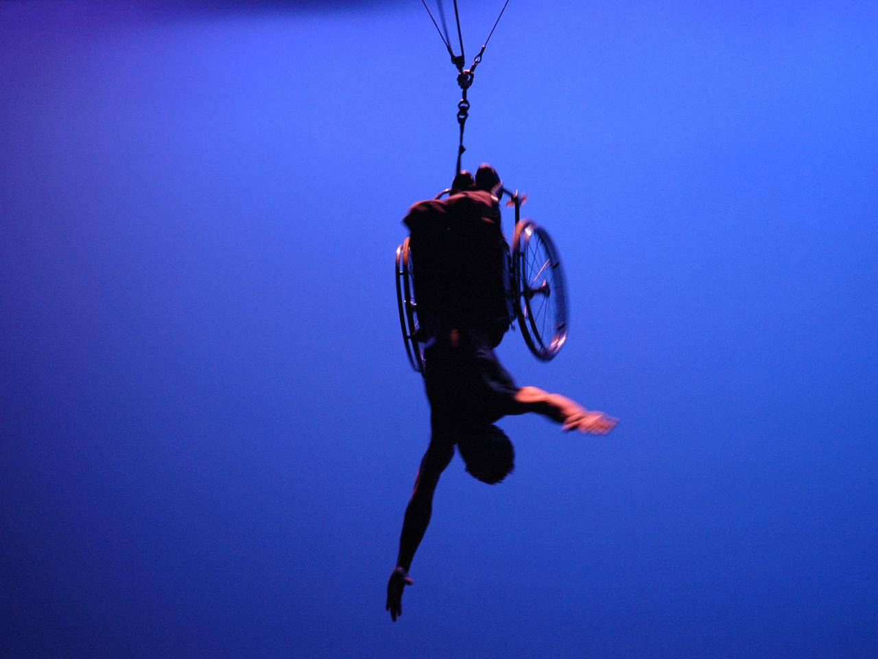 A person in a wheelchair suspended upside down with their arms extended, against a bright blue background.