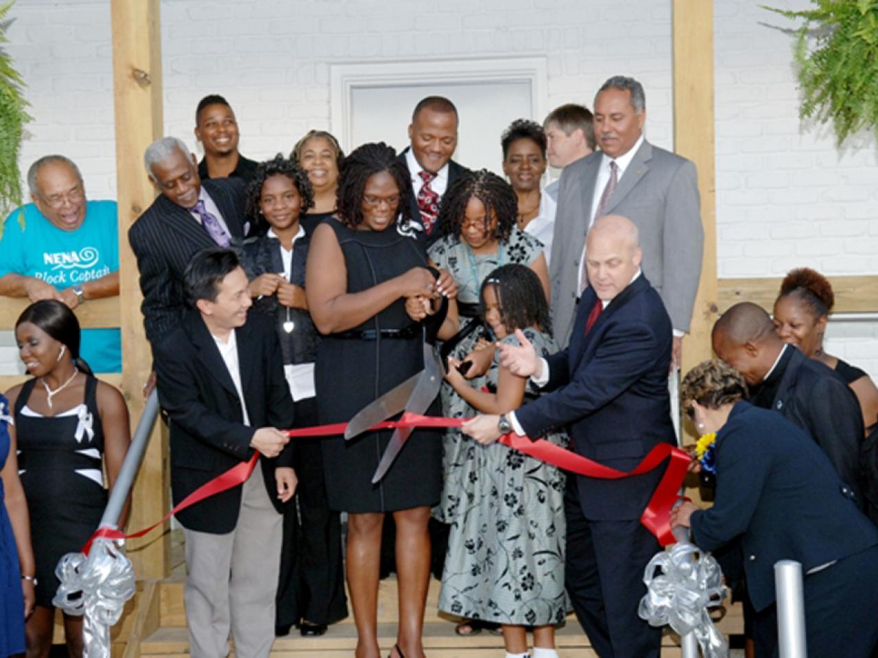 A group of mostly African-Americans of various ages gather on a stairway outside a building while two people hold scissors and are about to cut a red ribbon.