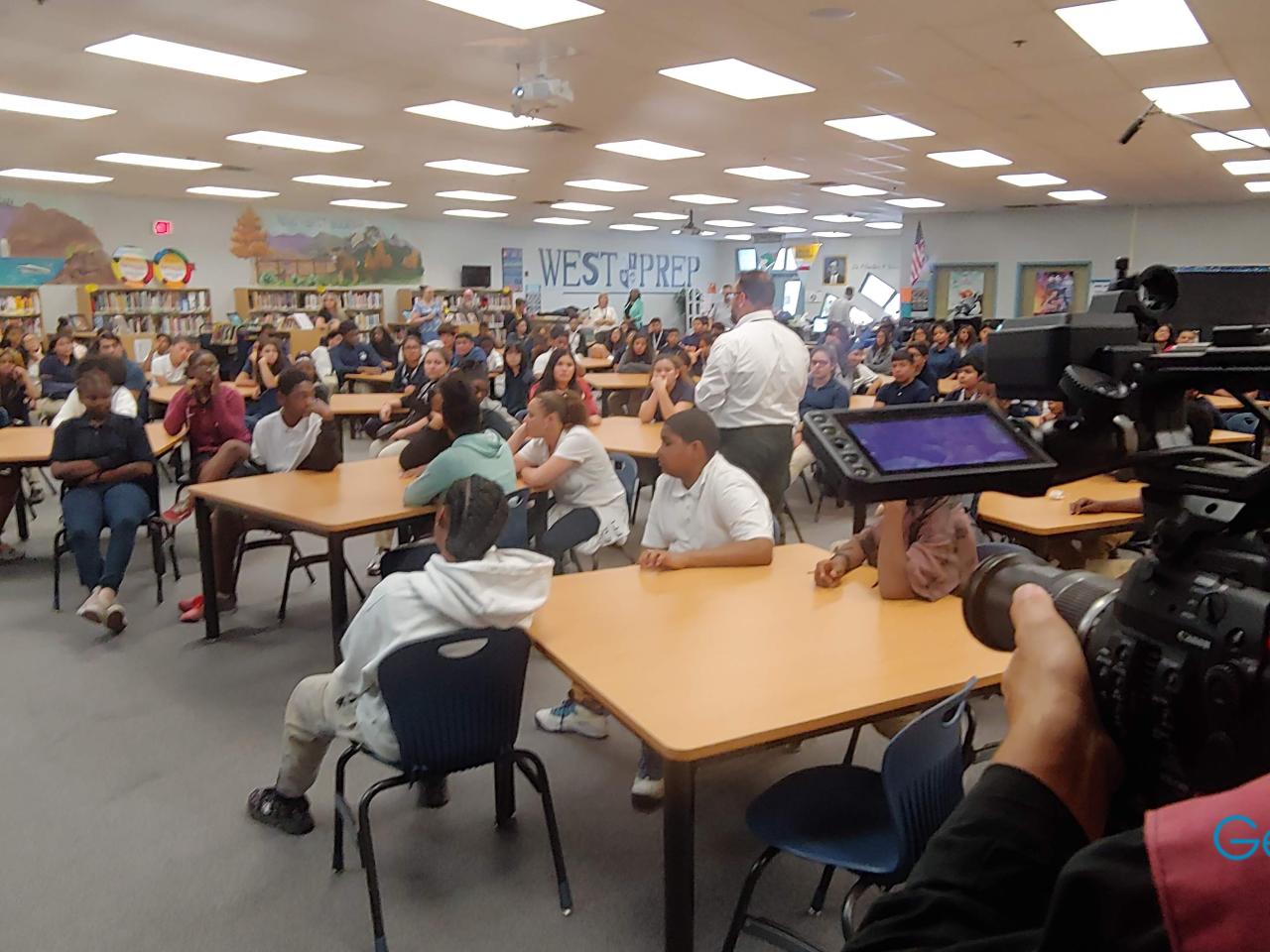 The “Trust Me” cinematographer focuses in as he films a library or lecture hall full of West Prep middle or high school students sitting in groups at small tables. The students are a mix of genders and ethnicities. Many are focused on an administrator who walks through the room, speaking to them.