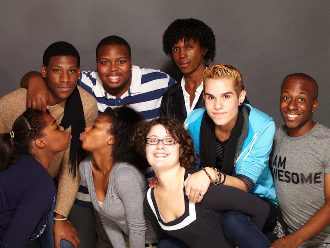 A group of queer teenagers having fun together. They are mostly Black youth. Two girls pucker up to kiss. The boys behind them have their arms around each other. One kid rides on another girl’s back. One kid wears a t-shirt that says I Am Awesome.