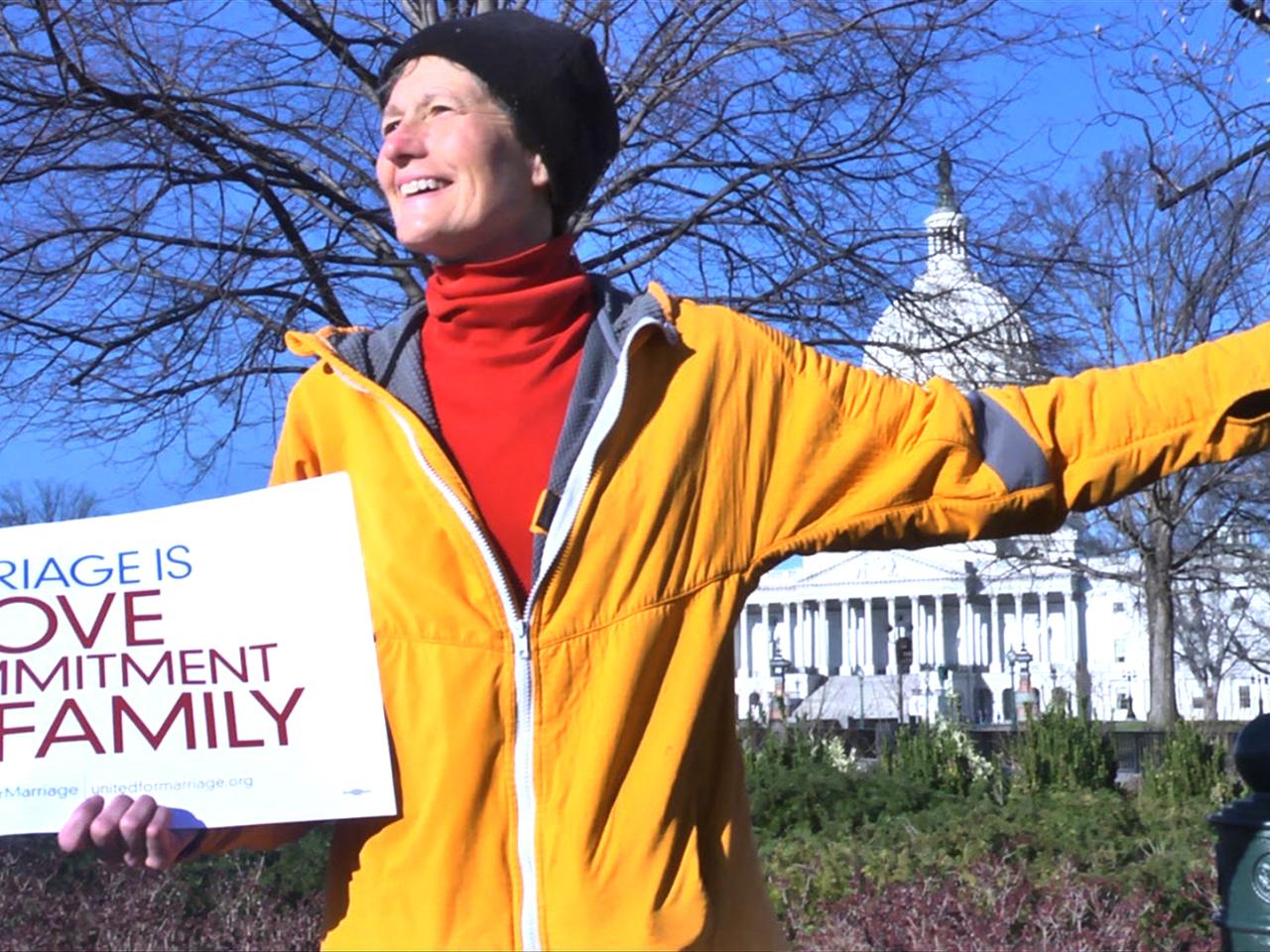 A white woman stands outside in front of a bare tree with blue sky. She smiles broadly, looking to the side. She is holding a white sign that reads “Marriage is Love, Commitment, Family” in blue and purple text. She wears a bright yellow zip up jacket which is half-unzipped to reveal a bright red turtleneck underneath and a black beanie on her head.