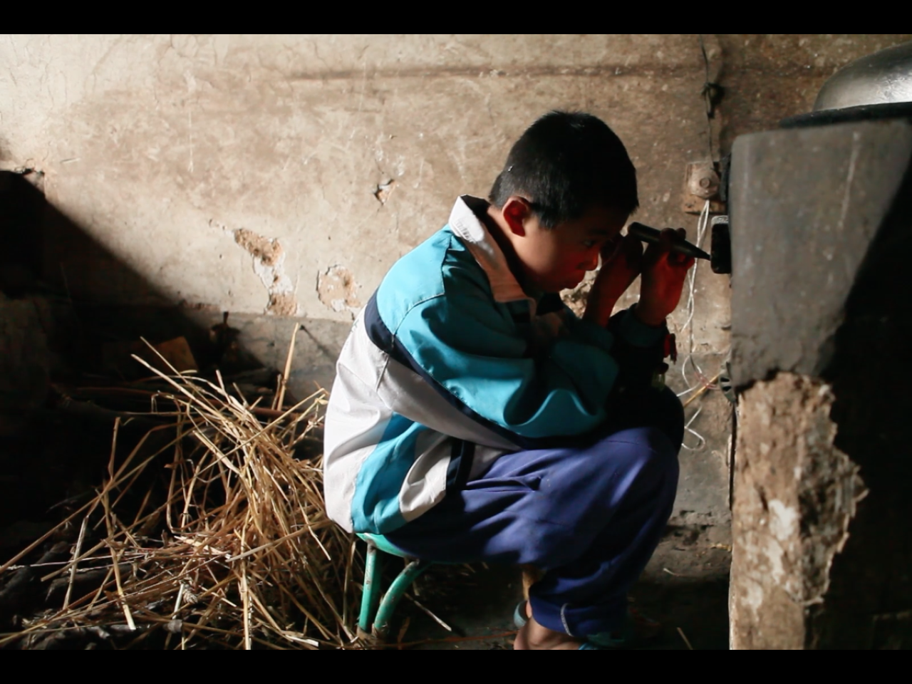 A young Chinese boy, maybe ten years old, sits on a low stool, working intensively on an electrical device attached to an old decaying structure.