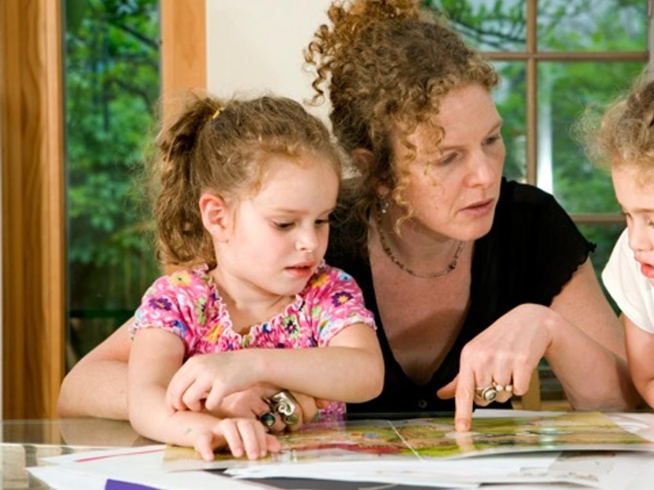 A still image from Sperm Donor X. Filmmaker Deirdre Fishel, a white woman with curly red hair pulled back, sits at the kitchen table reading a book with two young girls with curly red hair.