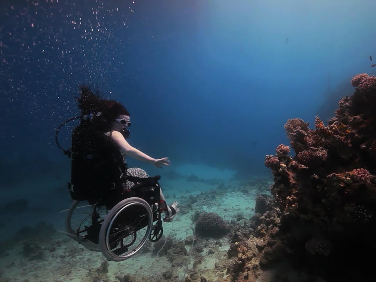 A still from the New Day film Fixed: The Science of Human Enhancement. A woman in a wheelchair is scuba diving. She floats slightly above the ocean floor. Her arms are bare and she does not appear to be wearing full scuba gear other than small goggles. Her hair drifts up in the water and she holds her arms out to the side for balance.