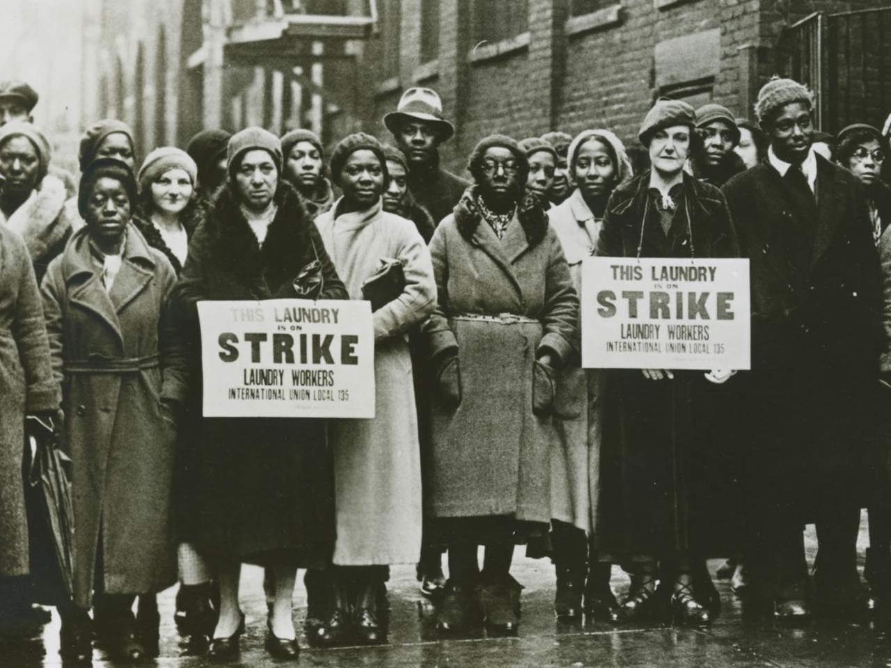 A black and white photo taken in the 1930’s of a group of striking workers who are predominantly Black women. Two women hold signs that say This Laundry is on Strike. Laundry Workers International Union Local 135.